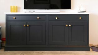 A sleek dark grey cabinet with gold knobs stands on a light wood floor. A TV sits on top, by a yellow vase and diffuser, creating a modern, chic vibe.