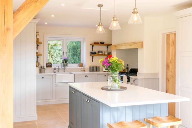 Rustic Shaker kitchen with exposed beams, grey cabinetry, island with two bar stools and a large vase of pink and yellows flowers.