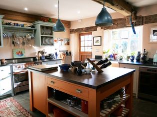 A warm, inviting kitchen features an orange island with cookware, under blue pendant lights. Rustic decor and natural light add a cozy, homely feel.