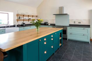 Spacious kitchen with teal cabinetry, wooden island, and modern appliances. Vase of flowers on the island, white tile backsplash, slate floor.