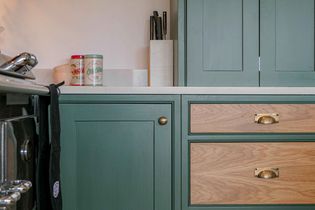 Stylish kitchen with sage green cabinets, wooden drawers, and brass handles. Countertop holds spice tins and a knife block.