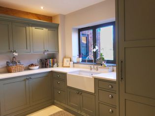 A cosy kitchen with light grey cabinets and white workrtops. Features a farmhouse sink, a window with plants, and warm, inviting lighting.