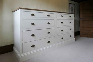 A large, white wooden dresser with multiple drawers and brass handles stands against a beige wall. It has a wooden top and exudes a classic, minimalist style.
