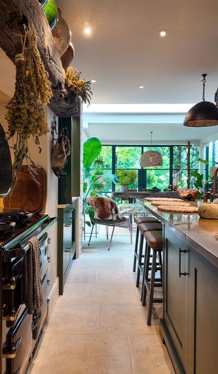 Warmly lit kitchen with the copper island and bar stools on the right, stove on the left. Dining table and greenery in the background.