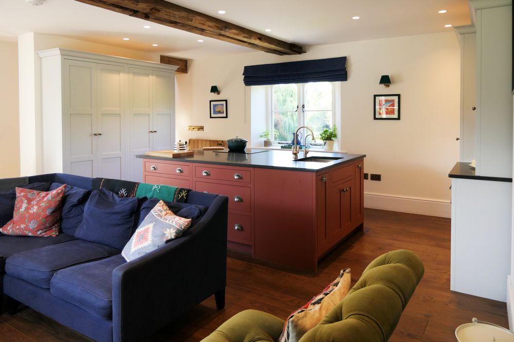 Bright open-plan Shaker kitchen and living room with a navy sofa, reddish-brown island, white cabinets, and a window overlooking greenery.