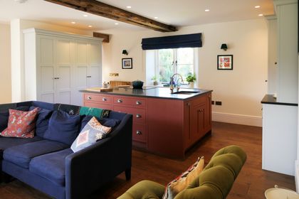 Bright open-plan Shaker kitchen and living room with a navy sofa, reddish-brown island, white cabinets, and a window overlooking greenery.