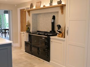 Stylish kitchen with a large black stove set in a marble alcove. Wooden shelves display small plants. A doorway leads to a bright dining room.