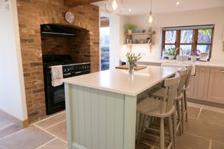 Shaker kitchen with light wood cabinets and a large island featuring bar stools. A brick arch houses a black oven. Shelves hold plants and decor.