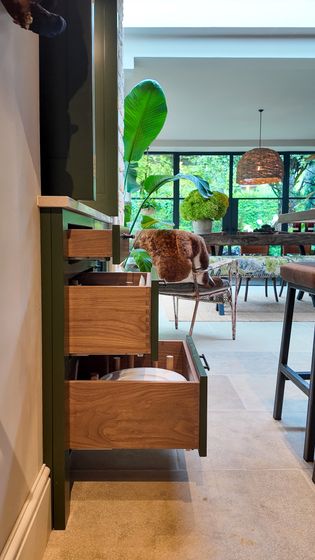 Warmly lit shaker kitchen with open dovetailed drawers, with the dining table and greenery in the background.