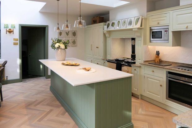 A neutral and sage green Shaker kitchen with a large floral arrangement in the centre of the island, pendant lighting and botanical artwork around the kitchen.
