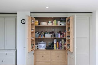 Open kitchen pantry with light wood shelves filled with spices, jars, and snacks. A bread container and cake box add a homely touch.
