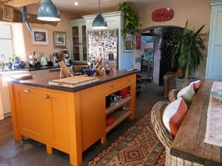 Warm, cozy kitchen with an orange island, woven wicker chairs, and blue cabinets. Rustic decor, colorful rugs, and leafy plants add charm and vibrancy.