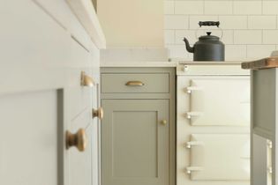 Modern kitchen close-up with cream cabinets, brass handles, cream AGA, and black kettle. Tile backsplash and light countertop add elegance.