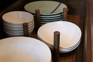 Stacked white and teal ceramic plates and bowls in a wooden drawer separated by wooden pegs, conveying an organized and minimalist kitchen vibe.