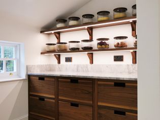 Shaker kitchen pantry with wooden cabinets, marble countertop, and shelves holding glass jars filled with grains, dried fruits, and eggs. Cozy and organized.