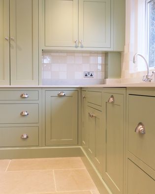 Sage green kitchen cabinets with silver cup handles, beige tile backsplash, and neutral stone floor create a calm, elegant atmosphere.
