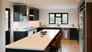 Shaker kitchen with dark cabinets, a long white island, and orange bar stools. Stainless steel appliances complement the sleek design. Natural light filters through a large window, creating a bright, inviting atmosphere.