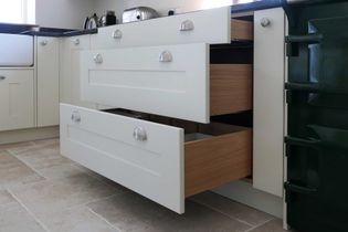 Open kitchen drawers with silver handles wood interiors. Light-coloured cabinets and tiled floor create a clean, modern ambiance.