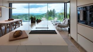 Modern kitchen with a large island, featuring a countertop stove, a loaf of bread on a cutting board, and panoramic views of nature through large windows.