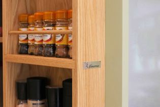 Wooden spice rack with neatly organized glass jars of spices, including cloves and cinnamon, each with an orange lid. A label reads "Minerva" Cosy kitchen vibe.