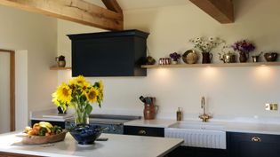 A cozy kitchen with a wooden beam ceiling features a vase of sunflowers on a white island. Dark cabinets, a farmhouse sink, and a shelf with assorted pottery add charm.