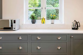 Shaker kitchen with grey drawers, shiny silver handles, a white quartz worktop, and a window with potted plants. A microwave and kettle add practicality.