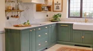 A Shaker kitchen with green cabinetry, a large farmhouse sink, and a quartz worktop, featuring wooden shelves with kitchen utensils and decorations.