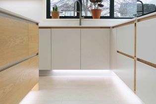 Modern kitchen with sleek white cabinets and wooden accents. Potted plants sit on the worktop, while under-cabinet lighting brightens the floor.