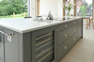 Shaker kitchen with a gray island featuring drawers and a glass-front wine fridge. The white countertop holds a kettle and vase with flowers. Large windows provide natural light, creating a serene ambiance.