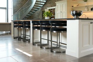 Large kitchen island with bar stools running alongside it. Curved staircase in the background with a white cabinet behind.