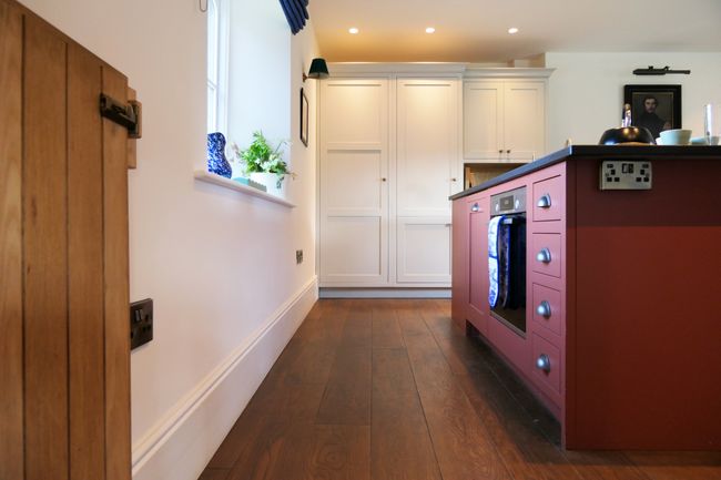 Warmly lit Shaker kitchen with cream cabinets, a red-brown island, wooden flooring, and a window with a potted plant.