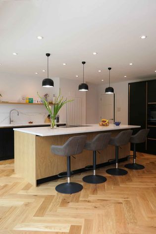Modern kitchen with a sleek island, white countertop, and four gray barstools. Pendant lights hang above. Warm wooden flooring and minimalist decor create a cozy feel.