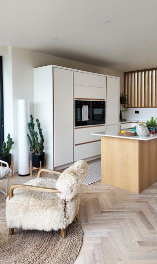A modern kitchen with white cabinetry, a wooden accent wall, and a central island. There are plants and kitchen appliances visible, along with a soft, cosy chair in the foreground.