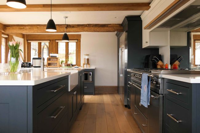 Spacious kitchen with dark cabinets, white worktops, and wooden floors. Natural light streams in through large windows, creating a warm, inviting atmosphere.
