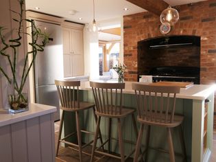 A shaker kitchen features a green blue island with three wooden chairs, a stainless steel fridge, brick oven, bamboo plant, and hanging bulb lighting. Warm and inviting atmosphere.