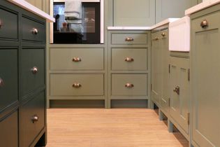 Light green kitchen cabinets with brass handles and a white farmhouse sink create a warm, rustic feel. Light wood flooring enhances the calming atmosphere.