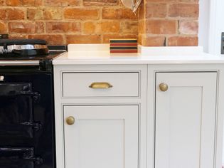 Elegant kitchen scene featuring a vintage dark stove beside light grey cabinets with brass handles, set against a rustic brick wall and white countertop.