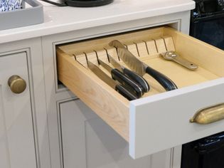 Open kitchen drawer with wooden dividers, holding four knives with black handles and a metal bottle opener. Bright, organized setting.
