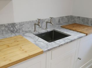 Light worktop with dual taps over a metal sink between two wooden cutting boards. The setting is a clean, modern kitchen.