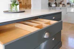 Open kitchen drawer with wooden dividers in a shaker kitchen island. Smooth blue cabinetry contrasts with a light countertop. 