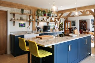Spacious kitchen with blue cabinets and a large marble island. Yellow bar stools add a pop of color. Shelves display plants and decor, creating a cozy vibe.