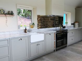 Shaker kitchen with grey cabinets, stone backsplash, and granite countertops. Features a farmhouse sink, stainless steel oven, and bright windows.