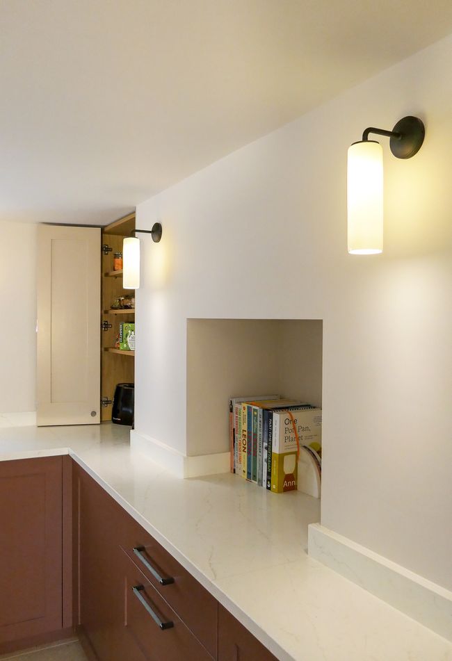 A Shaker kitchen with a quartz countertop, wall-mounted light fixtures, and open shelving displaying books. A countertop cabinet door is partially open in the background.