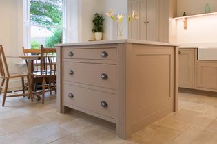 A shaker kitchen features a beige island with three drawers, sleek handles, and a flower vase. Nearby, a table with wooden chairs sits by a window.