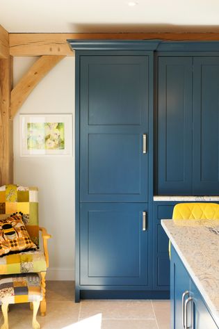 A cozy kitchen corner featuring a large blue cabinet, a patchwork quilted chair with a yellow cushion, and a wooden beam. Warm and inviting ambiance.