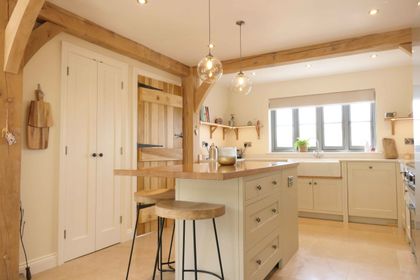 Bright kitchen with wooden beams, an island with two stools, pendant lights, cream cabinets, and large window. A rustic and inviting ambiance.