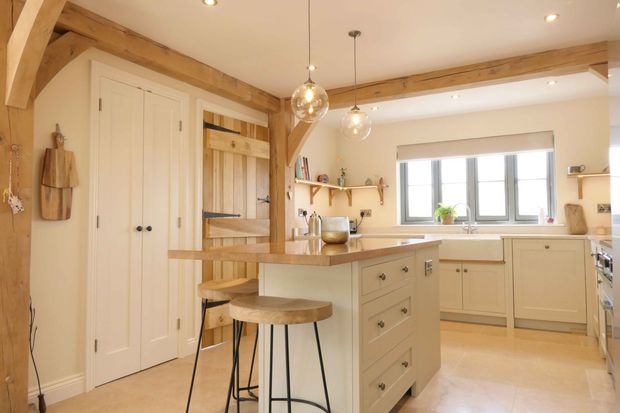 Bright kitchen with wooden beams, an island with two stools, pendant lights, cream cabinets, and large window. A rustic and inviting ambiance.
