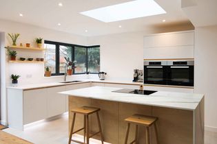 A modern kitchen with light wood and white cabinetry, a central island with stools, open shelves with plants, and corner windows for daylight. Bright and inviting.