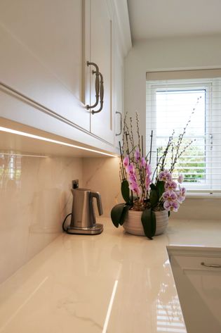 Shaker kitchen corner with sleek white cabinets and a polished marble countertop. A stainless steel kettle and blooming orchids add elegance near a window.