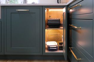 Open kitchen cabinet with green doors and brass handles, revealing stacked black cookware and neatly arranged baking trays. Warm lighting inside the cabinet.